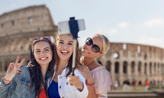 vacaciones de verano, vacaciones, viajes, tecnología y concepto de personas: grupo de mujeres jóvenes sonrientes que toman fotos con un teléfono inteligente en un palo selfie sobre el coliseo en el fondo de Roma