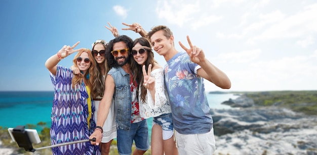 Foto vacaciones de verano, vacaciones, viajes, tecnología y concepto de personas - jóvenes amigos hippies sonrientes tomando fotos con un smartphone en un palo de selfie y mostrando un gesto de paz sobre el fondo de la playa