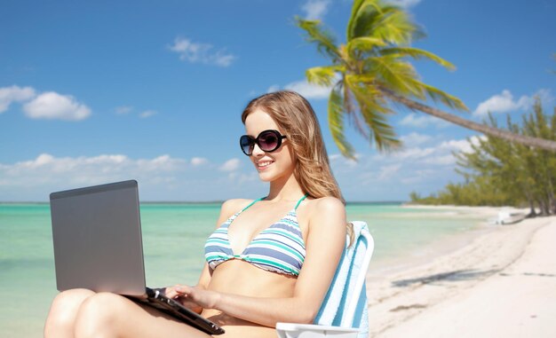 vacaciones de verano, vacaciones, viajes, tecnología y concepto de personas - mujeres sonrientes con gafas de sol con computadora portátil tomando el sol en el salón sobre una playa tropical exótica con fondo de palmera