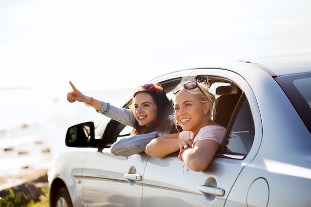 vacaciones de verano, vacaciones, viajes, viajes por carretera y concepto de personas: adolescentes felices o mujeres jóvenes sentadas en un camión a la orilla del mar y señalando con el dedo a algo