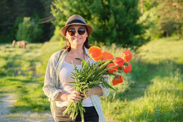 Vacaciones de verano, viaje por la naturaleza, hermosa mujer madura caminando por la carretera con ramo de flores de amapolas, fondo del atardecer paisaje escénico