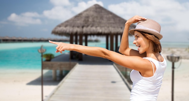 vacaciones de verano, viajes, gente y concepto de vacaciones - mujer joven feliz con sombrero señalando con el dedo sobre la exótica playa tropical con bungalow y fondo de la costa