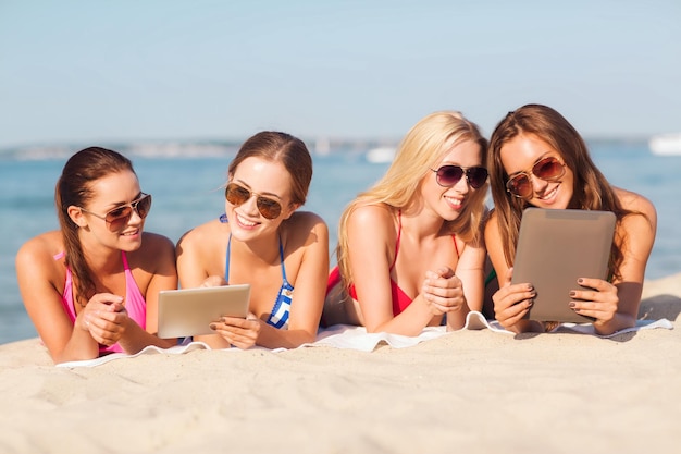vacaciones de verano, viajes, tecnología y concepto de personas - grupo de mujeres sonrientes en gafas de sol con computadoras de tablet pc tiradas en la playa