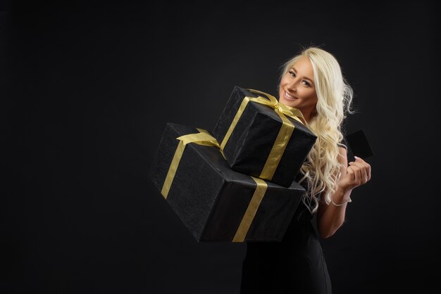 Vacaciones del viernes negro. Mujer rubia sosteniendo una tarjeta de crédito en la pared oscura. Cajas de regalo