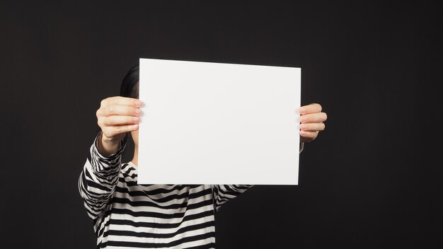 Vacía en blanco el papel A4 en manos de la mujer sobre fondo negro. La mujer usa camiseta de rayas blancas y negras.