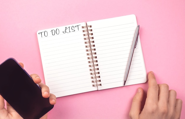 Vacía la lista de tareas pendientes para el año nuevo. Cuaderno de mesa rosa, fondo del concepto de planificación. Mano con smartphone y bolígrafo. Foto de vista superior