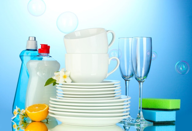 Foto vacíe platos limpios, vasos y tazas con esponjas líquidas para lavar platos y limón sobre fondo azul.