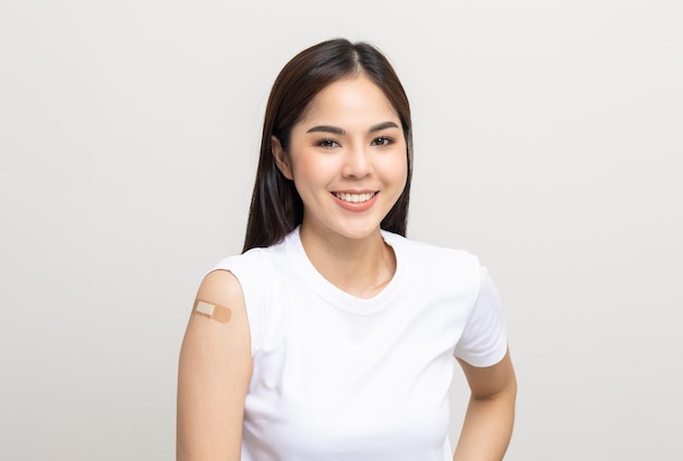 Vacinação. Jovem e bela mulher asiática recebendo uma vacina que protege o coronavírus. Sorrindo feliz fêmea mostrando o braço com curativo após receber a vacinação. Em fundo branco isolado.