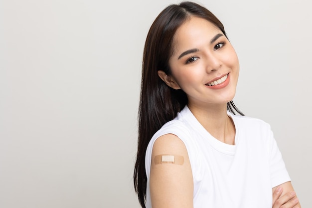Vacunación. Joven hermosa mujer asiática recibiendo una protección de vacuna contra el coronavirus. Sonriente mujer feliz mostrando el brazo con vendaje después de recibir la vacunación. Sobre fondo blanco aislado.