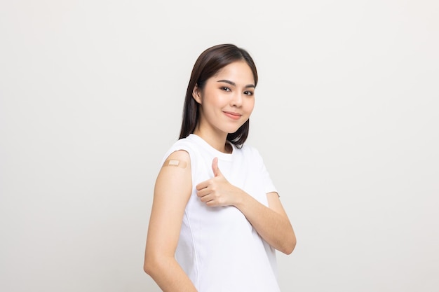 Vacunación. Joven hermosa mujer asiática recibiendo una protección de vacuna contra el coronavirus. Sonriente mujer feliz mostrando el brazo con vendaje y pulgares hacia arriba. Sobre fondo blanco aislado.