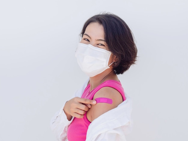 Vacunas, concepto de personas vacunadas. Las mujeres asiáticas felices con mascarilla y camisa sin mangas rosa señalando el vendaje en el propio hombro con una sonrisa después del tratamiento de vacunación sobre fondo blanco.