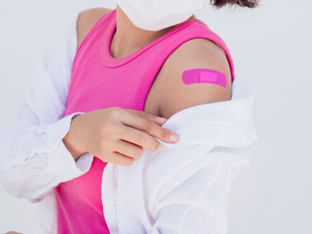 Foto vacunas, vendaje en concepto de brazo de personas vacunadas. vendaje rosa después del tratamiento de vacunación en el hombro de la mujer que usa mascarilla, camiseta rosa sin mangas y blanca sobre fondo blanco.