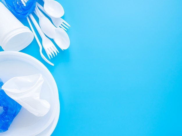 Vajilla desechable, cucharas de plástico blanco, tenedores, vasos y platos aislados sobre un fondo azul.