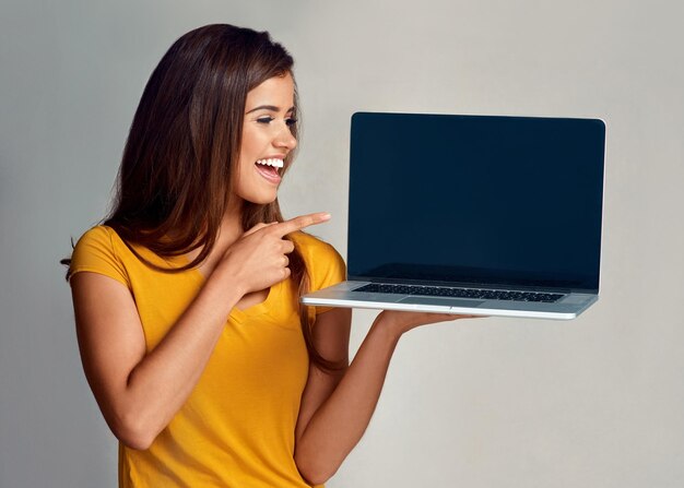 Valdrá la pena ver esto Foto de estudio de una mujer joven y atractiva que señala una computadora portátil con una pantalla en blanco contra un fondo gris