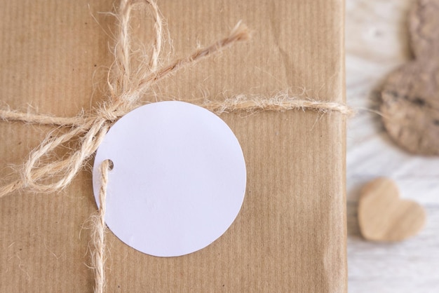 Valentins-Kraftpapier verpacktes Geschenk mit rundem Geschenkanhänger, der am Seilbogen in der Nähe der Draufsicht der Herzen hängt. Rustikale Frühlingsszene mit leerem Etikettenmodell, Kopierraum