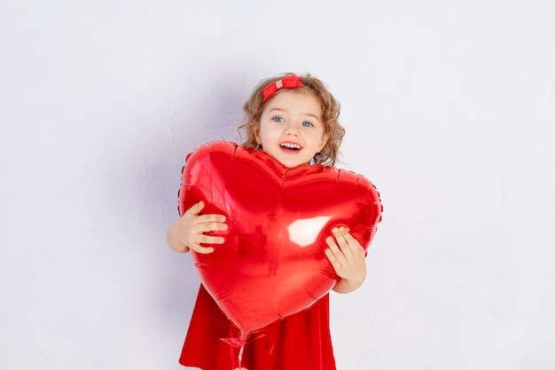 Valentinstag Baby. Ein kleines Mädchen in einem roten Kleid hält einen großen Ball in Form eines Herzens