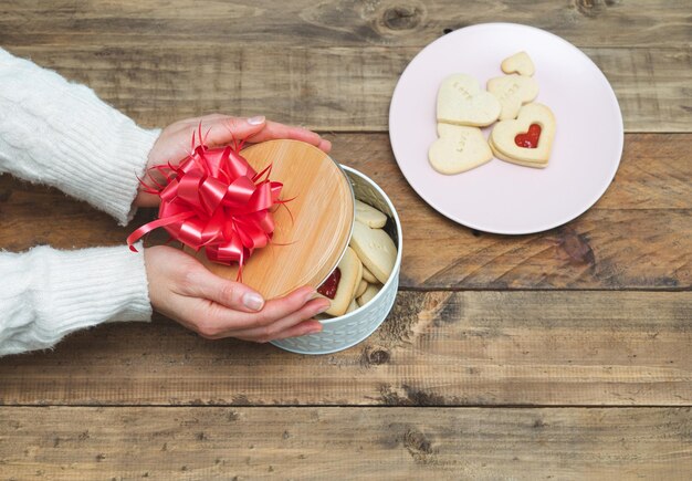 Valentinstag. Frauenhände, die Keksschachtel mit rotem Band und herzförmigen Keksen aufdecken. Platz kopieren.