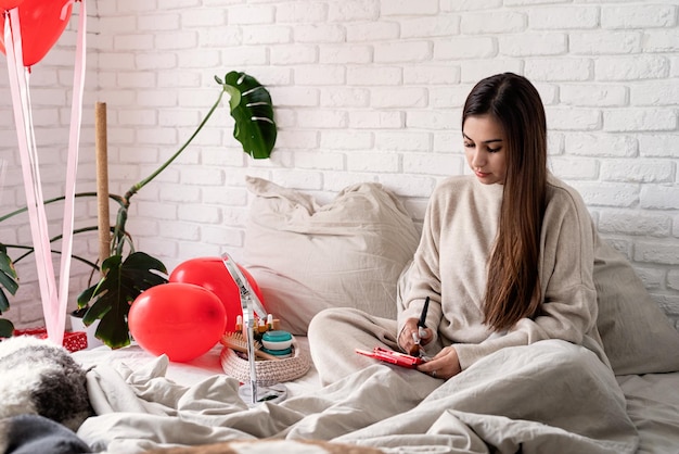 Valentinstag, Frauentag. Brünette junge Frau feiert Valentinstag, sitzt auf dem Bett und schminkt sich