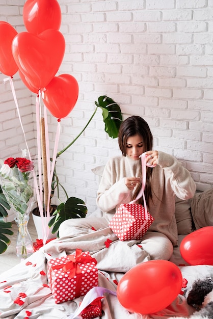 Valentinstag, Frauentag. Junge kaukasische brünette frau, die im bett sitzt und den valentinstag feiert, der die geschenke einwickelt