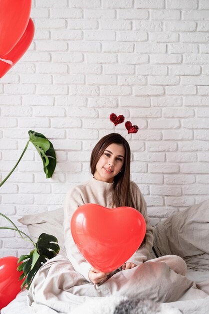 Valentinstag, Frauentag. Junge lustige Frau sitzt im Bett und feiert den Valentinstag mit Luftballons