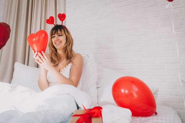 Valentinstag, Frauentag. Junges kaukasisches mädchen, das im bett sitzt und den valentinstag feiert, der einen herzförmigen ballon in ihren händen hält