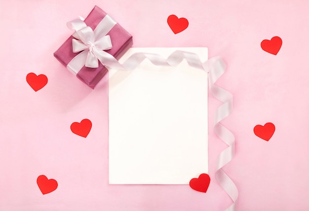 Foto valentinstag-grußkarte mit rosa geschenkbox, weißer schleife, langem gebogenem band und roten papierherzen