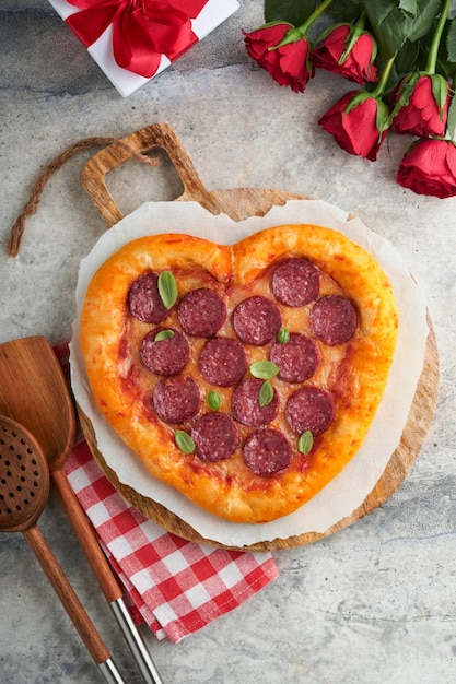 Valentinstag herzförmige Pizza mit Mozzarella Peperoni und Basilikum Weinflasche zwei Weinglas Geschenkbox auf hellgrauem Hintergrund Idee für ein romantisches Abendessen Valentinstag Draufsicht Mock-up