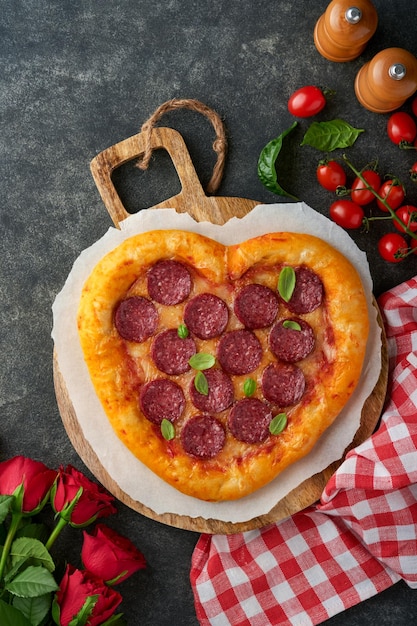 Valentinstag herzförmige Pizza mit Mozzarella Peperoni und Basilikum Weinflasche zwei Weinglas Geschenkbox auf schwarzem Hintergrund Idee für ein romantisches Abendessen Valentinstag Draufsicht Mock-up