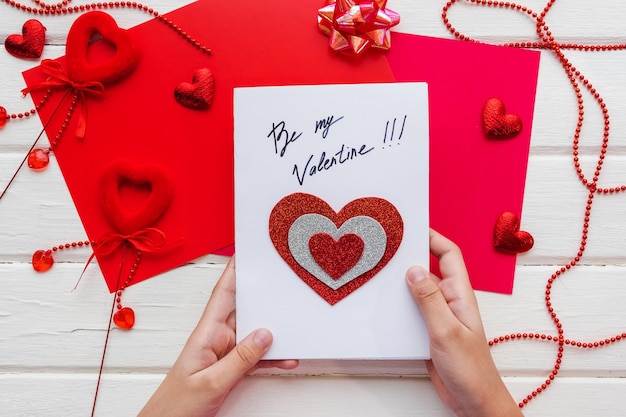 Valentinstag Hintergrund. Handgemachte Geschenk-Grußkarte zum Erstellen, Ausschneiden und Einfügen, Bastelpapier, Filz und DIY-Werkzeuge auf weißem Holz. Ansicht von oben, flach