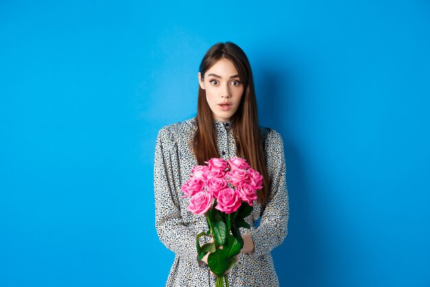 Valentinstag-Konzept. Überraschte Freundin, die einen schönen Blumenstrauß erhält und ungläubig in die Kamera schaut, auf blauem Hintergrund stehend.