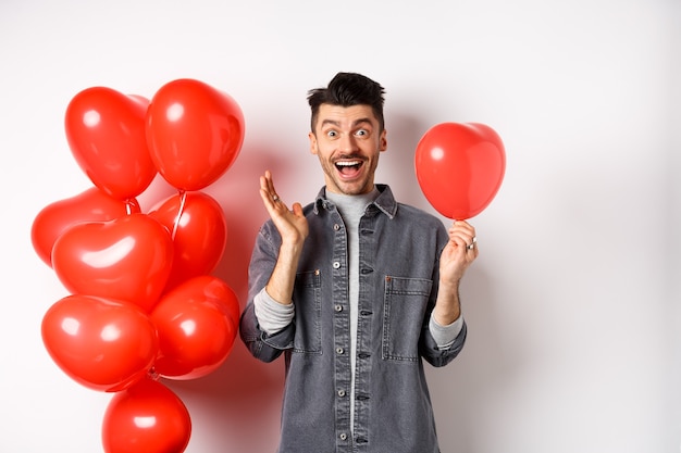 Valentinstag Konzept. Fröhlicher junger Mann, der Liebesfeiertag feiert, in der Nähe von roten Herzballons steht und überrascht aussieht, Freudenschrei, weiß.