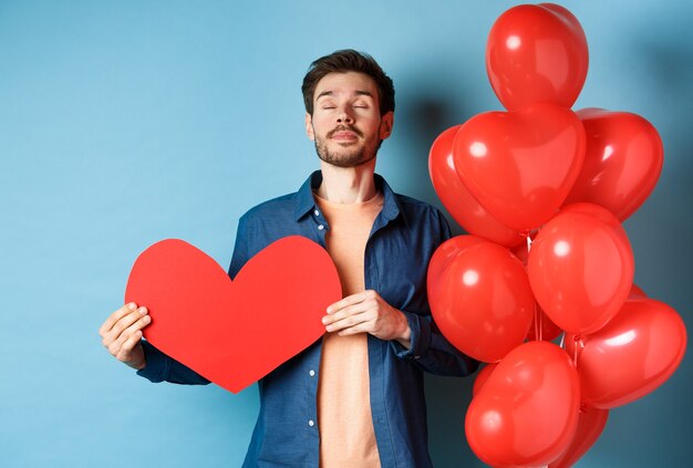 Valentinstag Konzept. Mann träumt von wahrer Liebe, hält roten Herzausschnitt und steht nahe romantischen Luftballons, blauer Hintergrund.