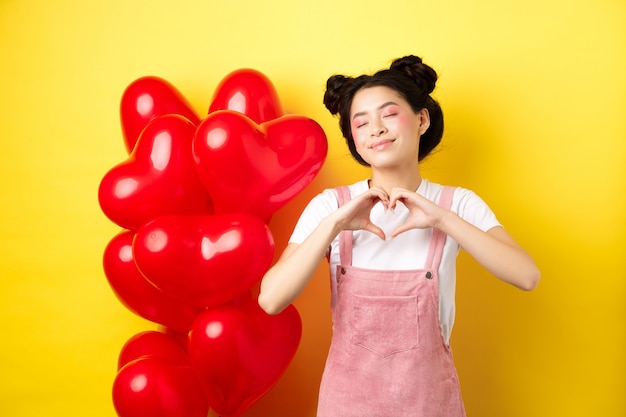 Valentinstag Konzept. Nettes asiatisches Mädchen, das von Romantik träumt, Augen schließt und Herzgeste zeigt, glücklich lächelnd, nahe roten romantischen Luftballons stehend, gelber Hintergrund steht.