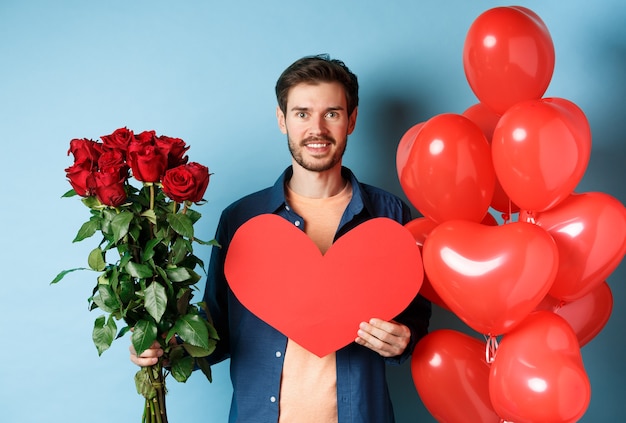 Valentinstag Romantik. Freund mit Bouquet von roten Rosen und Herzballons lächelnd, bringen Geschenke für Liebhaber am Valentinstag, stehend über blauem Hintergrund.