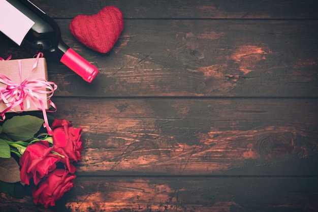 Valentinstag romantische Grußkarte. Rote Rosenblüten, Weinflasche, Geschenkbox und dekoratives Herz auf Holztisch. Freiraum.