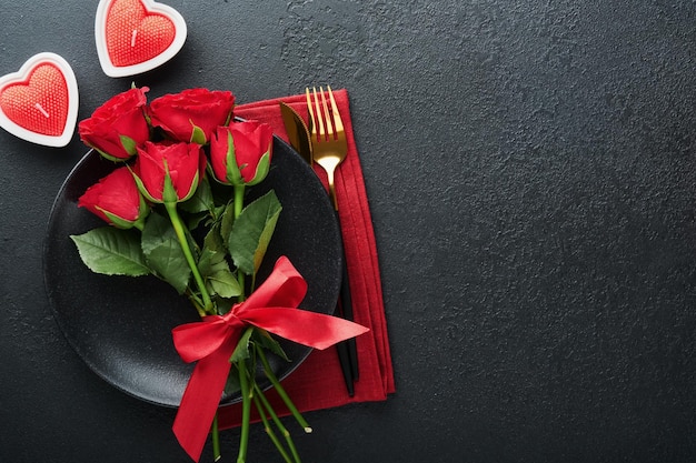 Valentinstag romantische Tischdekoration Leere Nahaufnahme von roten Rosen Wein Kerzen Abendessen schwarze Platte Messer Gabel und dekorative Seidenherzen auf schwarzem Hintergrund Urlaubskonzept Kopierbereich Ansicht von oben