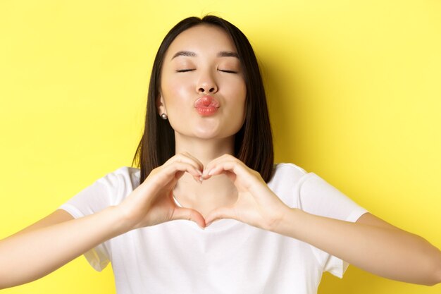 Valentinstag und Frauenkonzept. Schließen Sie herauf des hübschen asiatischen Mädchens im weißen T-Shirt, das lächelt und Herz zeigt, ich liebe Sie Geste, die über gelbem Hintergrund steht.