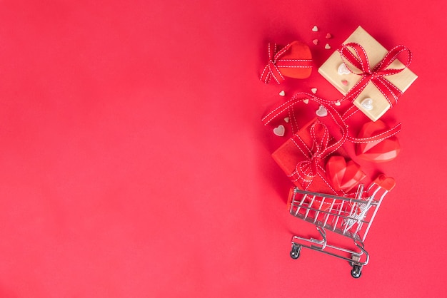 Valentinstag-Verkauf, Stillleben-Zusammensetzung mit festlicher Geschenkbox, rotem Herzen und Einkaufswagen