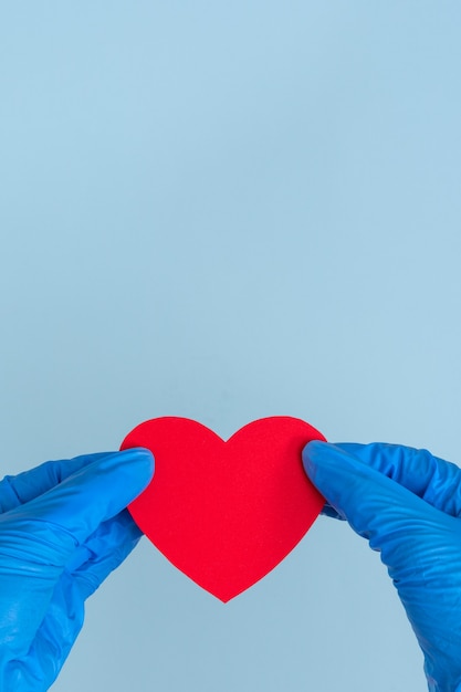 Valentinstag während der Coronavirus-Pandemie. Zwei blaue Latexhandschuhhände halten eine rote Herzform auf einem blauen Hintergrund, Nahaufnahme, Kopierraum, vertikaler Rahmen