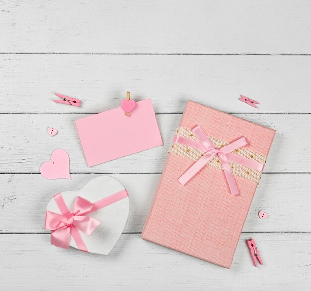 Valentinstagschablone von rosa Geschenkboxen mit Bandchiffonschleifen und Papiernotiz auf weißem hölzernem Tischhintergrund, schließen oben flaches Legen, erhöhte Draufsicht, direkt oben