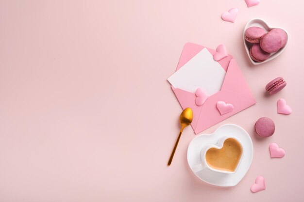 Valentinstagskarte. Rosa leerer Umschlag, Macaron-Makronenkeks und herzförmige Kaffeetasse auf rosa Hintergrund. 8. März, Valentinstag, Geburtstag der Mutter der Frauen. Flache Lage, Draufsicht, Kopierbereich.