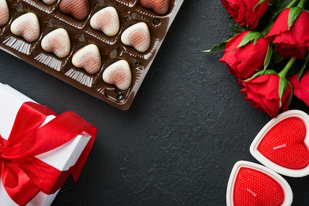 Valentinstagskarte Rosenrote Blumen, Brownie-Kekse und Pralinen auf herzförmiger Geschenkbox mit Wein auf schwarzem Hintergrund Grußkarte zum Mutter- oder Frauentag Draufsicht flach gelegt