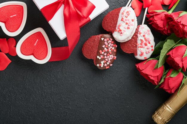 Valentinstagskarte Rosenrote Blumen, Brownie-Kekse und Pralinen auf herzförmiger Geschenkbox mit Wein auf schwarzem Hintergrund Grußkarte zum Mutter- oder Frauentag Draufsicht flach gelegt