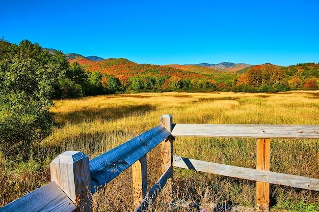 Valla de madera junto a grandes campos amarillos con coloridas montañas de otoño en la distancia