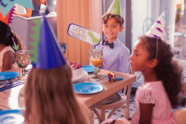 Vamos festejar. Menino alegre e relaxado usando chapéu de festa e segurando uma placa permite a festa com o dinossauro verde