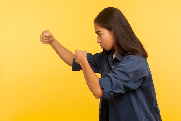 Vamos lutar. Vista lateral da menina corajosa confiante na camisa jeans, mantendo os punhos cerrados, boxe e socos para o lado, luta e conceito de autodefesa. tiro de estúdio isolado em fundo amarelo