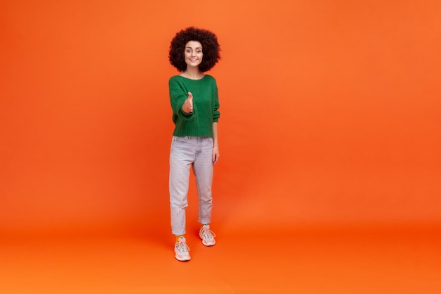 Vamos nos familiarizar Comprimento total da mulher com penteado afro vestindo suéter verde dando a mão, dando as boas-vindas, oferecendo aperto de mão, mostrando acordo. Tiro de estúdio interior isolado em fundo laranja.