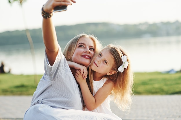 Vamos tirar uma selfie. Foto de uma jovem mãe e sua filha se divertindo na grama verde com lago ao fundo.