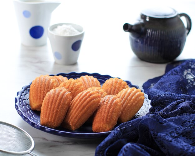 Vanille-Zitrone Madeleine auf blauer Keramikplatte. Berühmtes französisches süßes Muschelgebäck mit Zuckerstaub, serviert mit Tee. Perfekt für Tee- oder Kaffeezeit