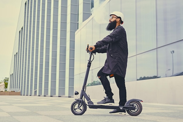 Vaping masculino hipster barbudo e andando de scooter elétrico sobre o edifício moderno.